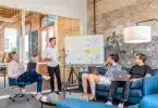 three men sitting while using laptops and watching man beside whiteboard